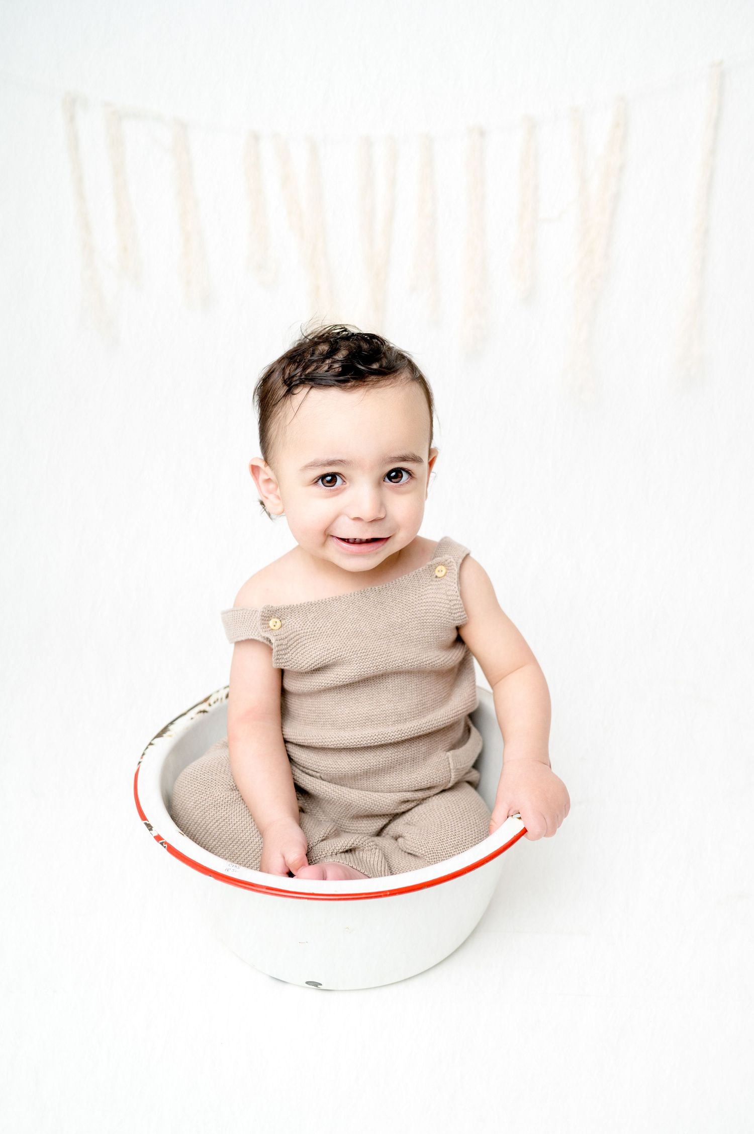Baby grins while sitting in white and red bowl wearing neutral colored outfit.