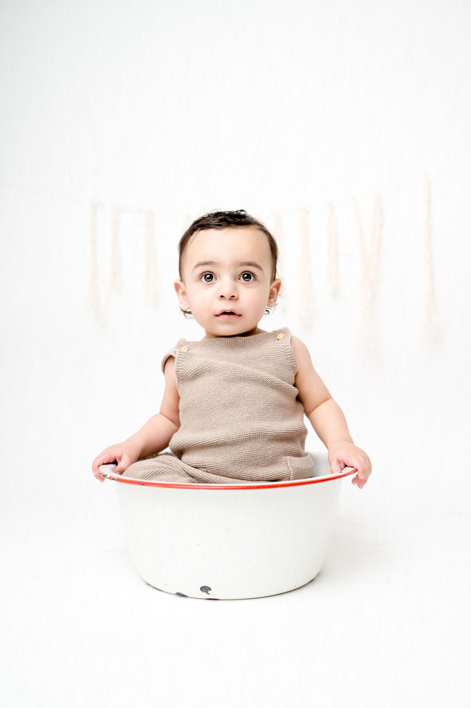 Baby in tan sleeveless outfit sits inside white bowl against plain background.