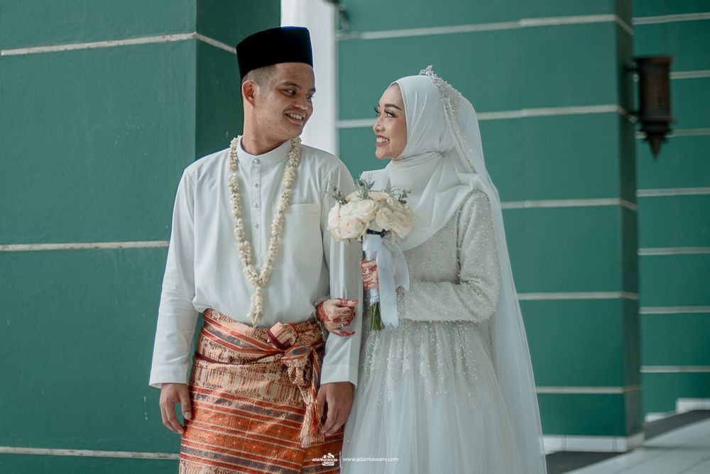 Muslim couple in traditional white wedding attire poses together against turquoise wall with floral bouquet.