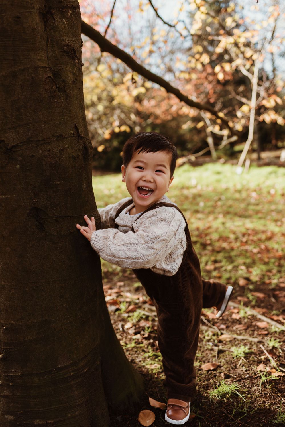 Toddler in brown overalls laughing while playing near tree trunk in autumn setting.