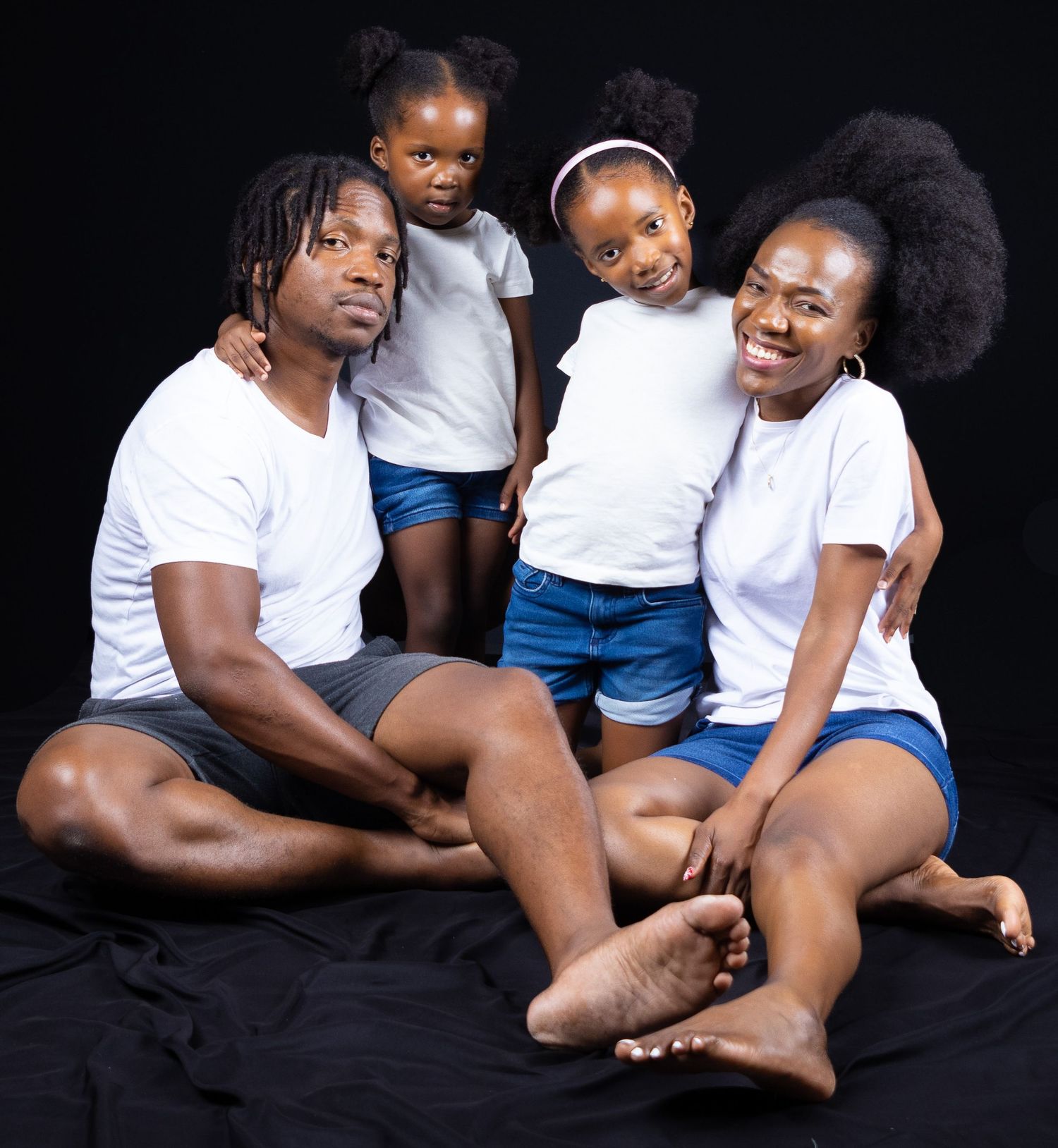 Family wearing matching white t-shirts and blue shorts pose together on black background.