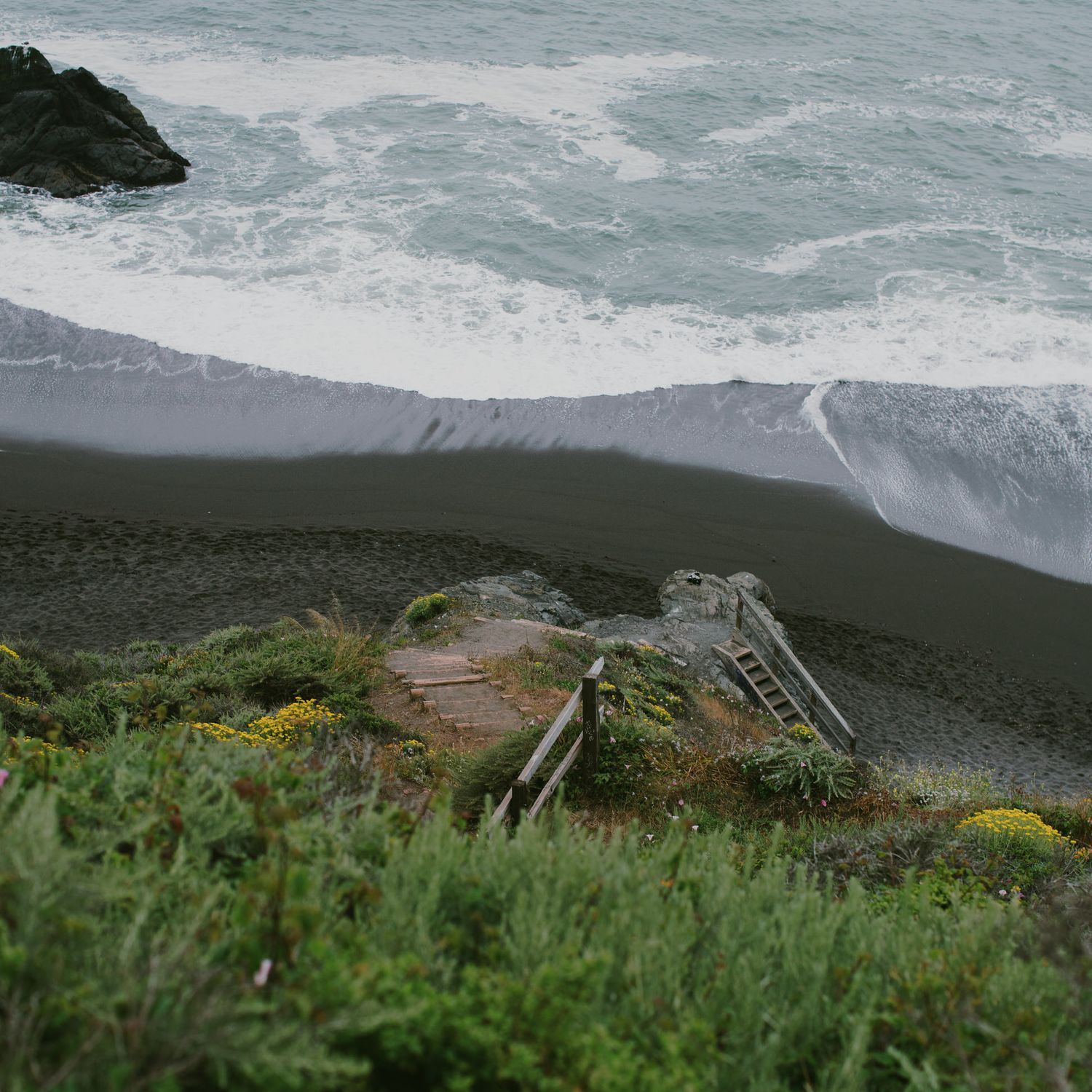 Dramatic black sand beach with crashing waves and wooden stairs leading down the coastal cliffside.