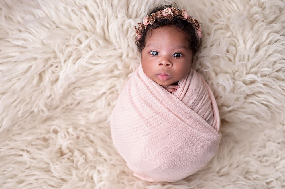 Newborn baby wrapped in pink swaddle with floral crown poses on fluffy white rug.