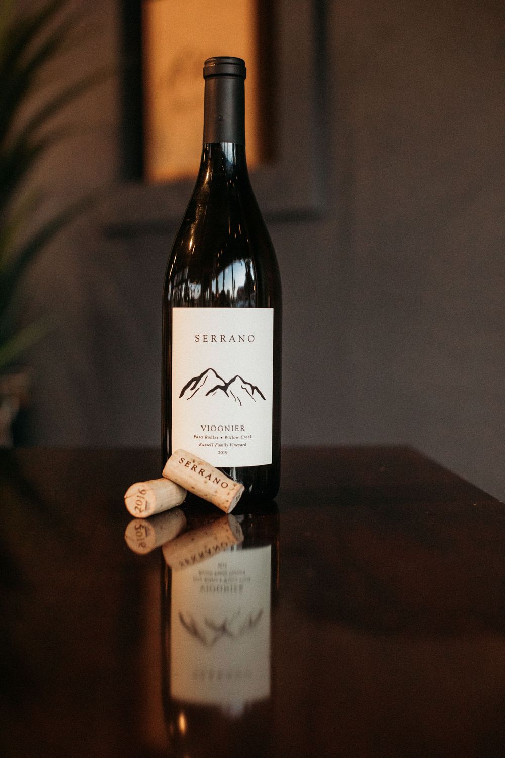 A bottle of Serrano wine with mountain logo displayed with corks on reflective surface.