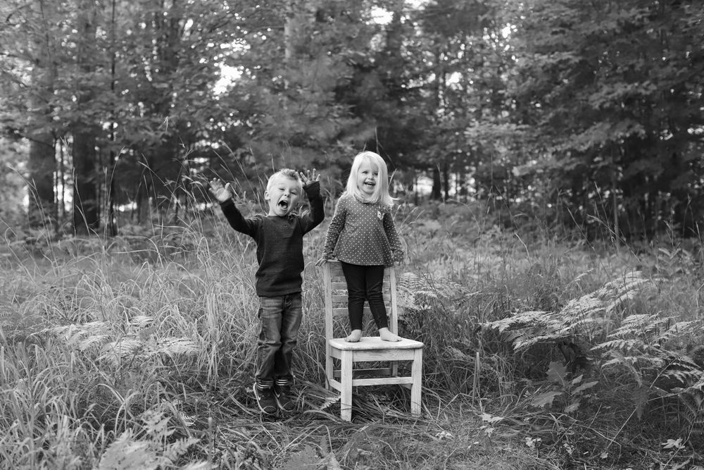 Black and white photo of children standing on white chair in forest clearing.