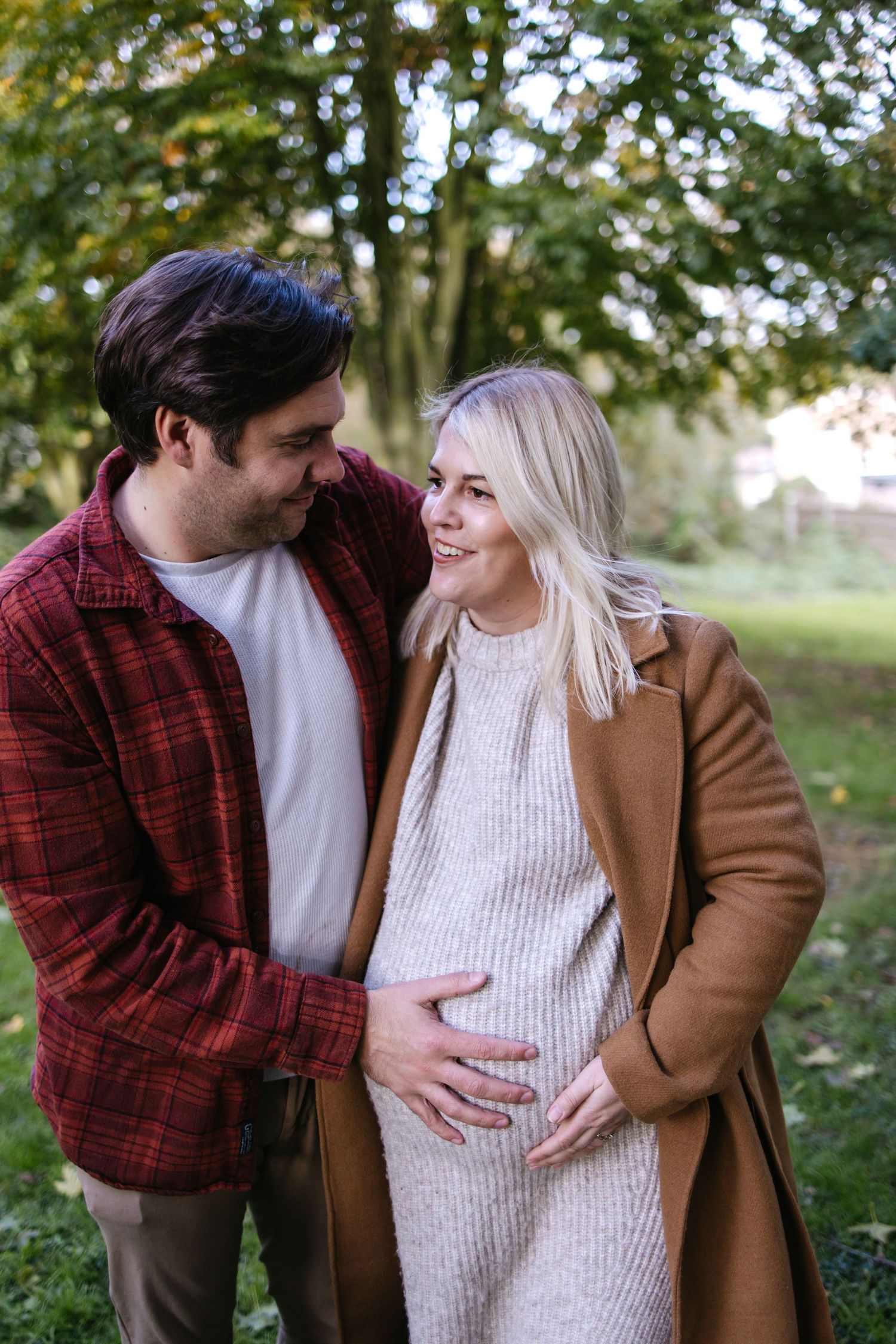 A couple in autumn attire share an intimate moment outdoors as they celebrate an expected arrival.