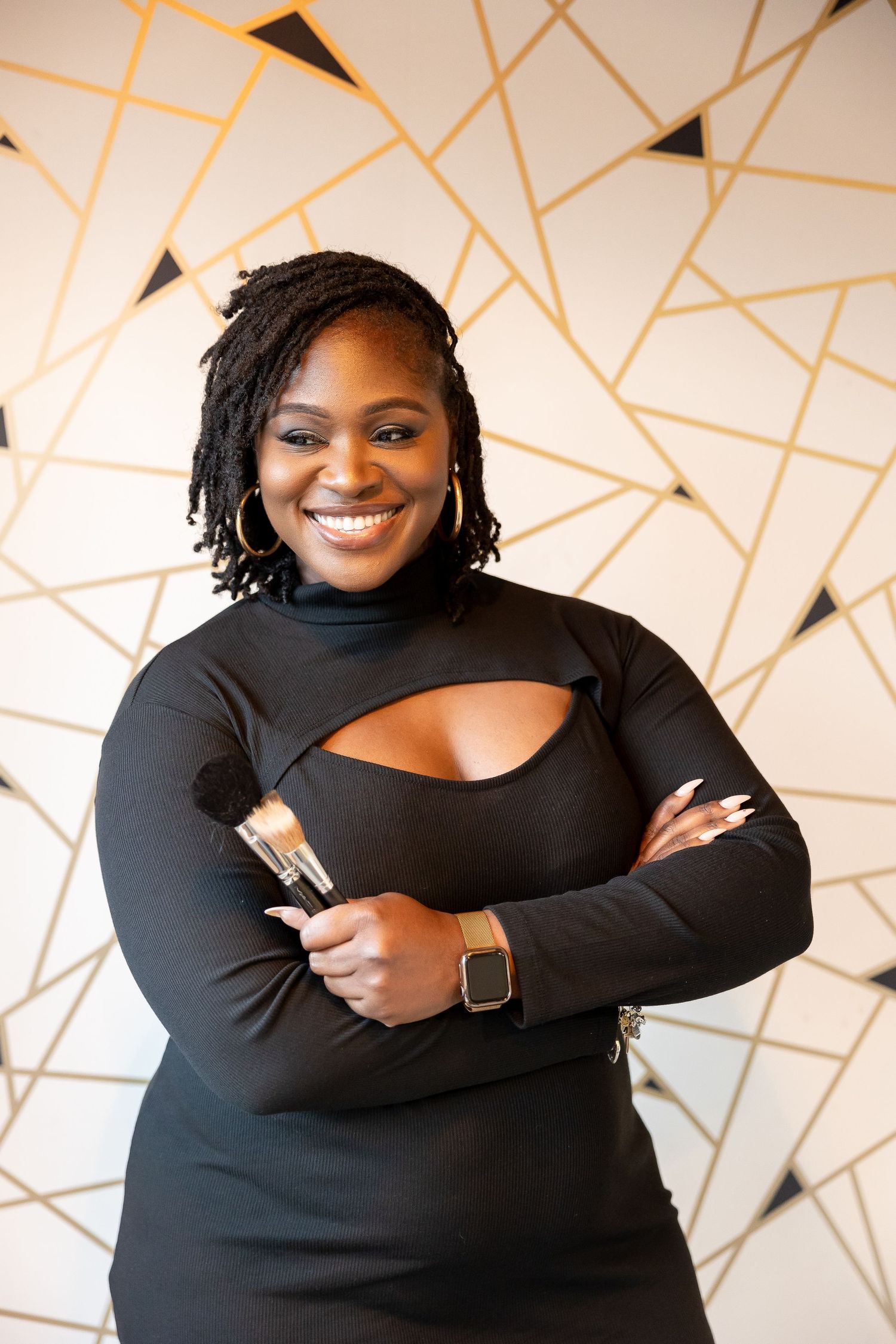 Smiling makeup artist in black dress holding brushes, posing in front of geometric gold and white wall.