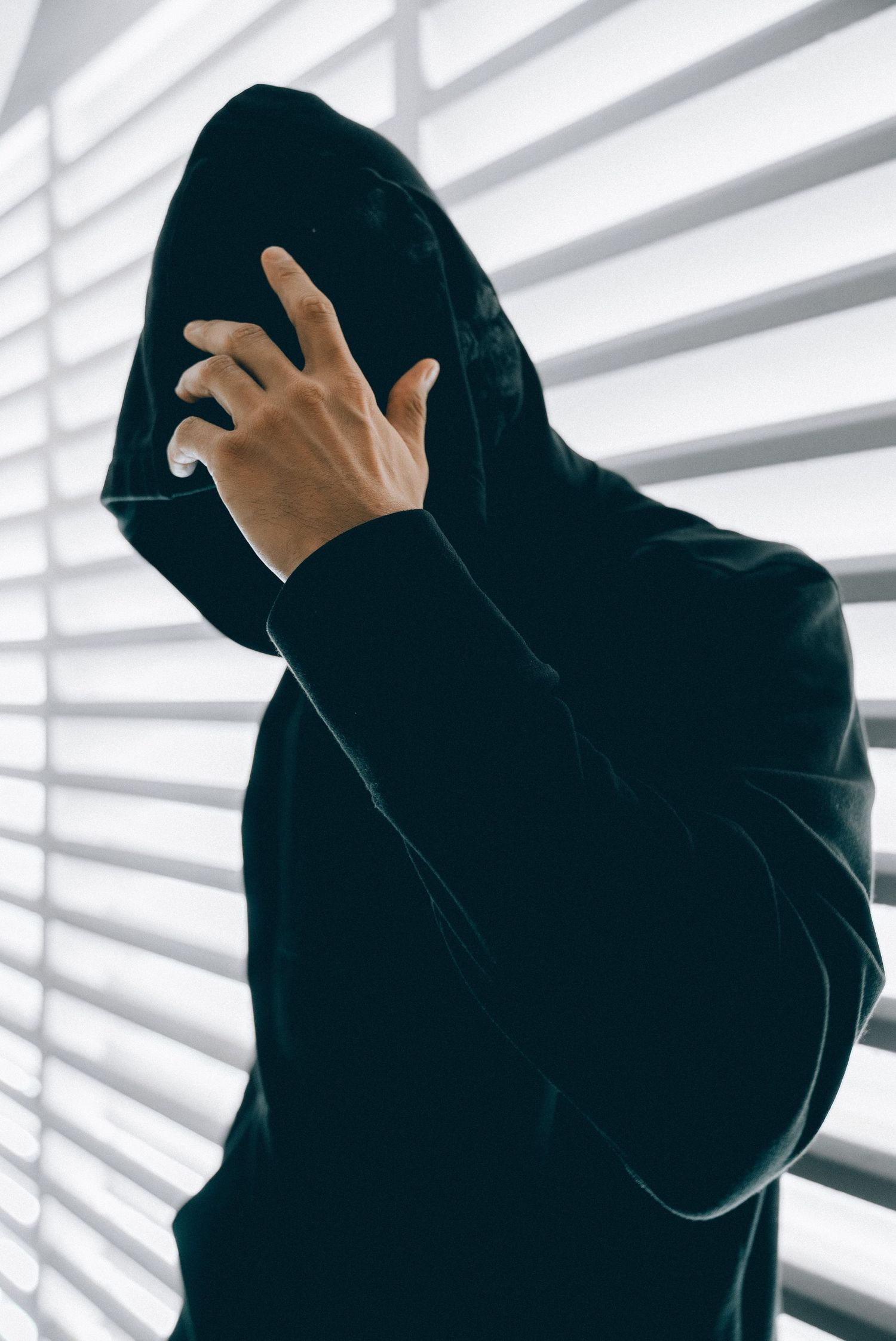 Silhouetted figure in black hoodie covering face with hand against window blinds in dramatic lighting.