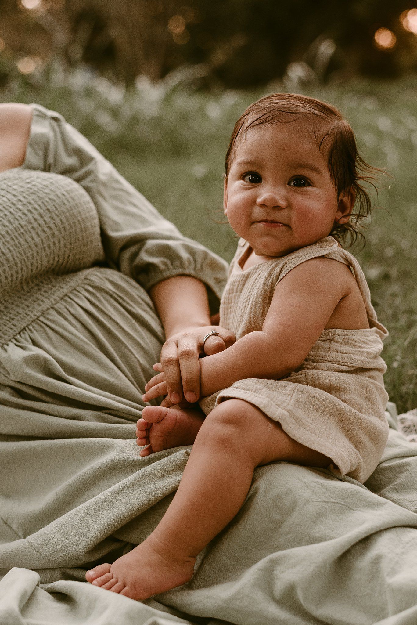 A small child in a beige dress sits on soft gray fabric with a joyful expression at sunset.