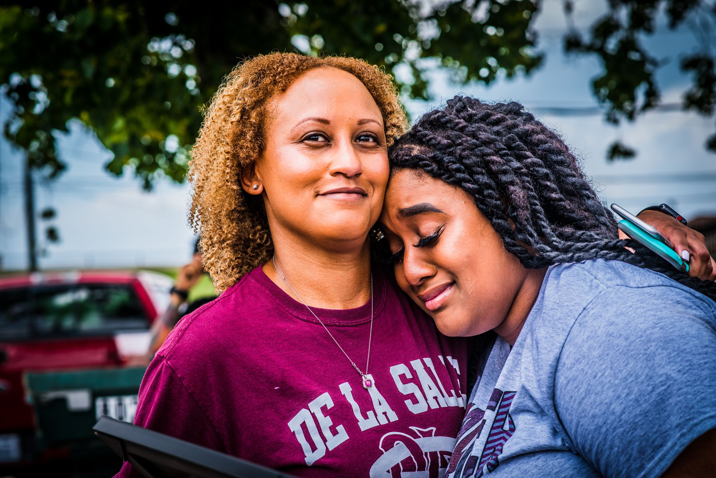 Two friends share an emotional moment outdoors, one wearing a burgundy sweatshirt.