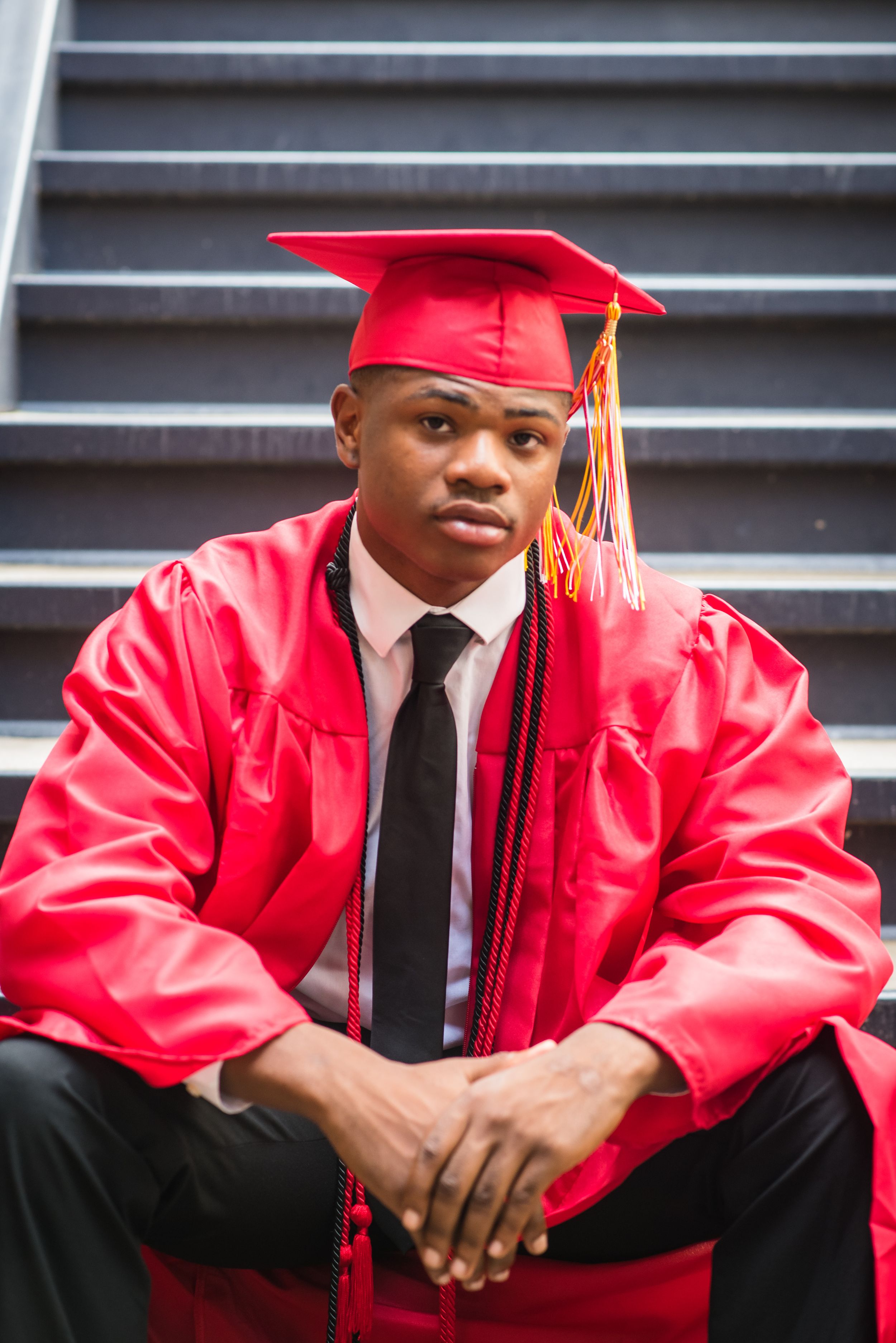Graduate in red cap and gown sits confidently on stairs for graduation photos.