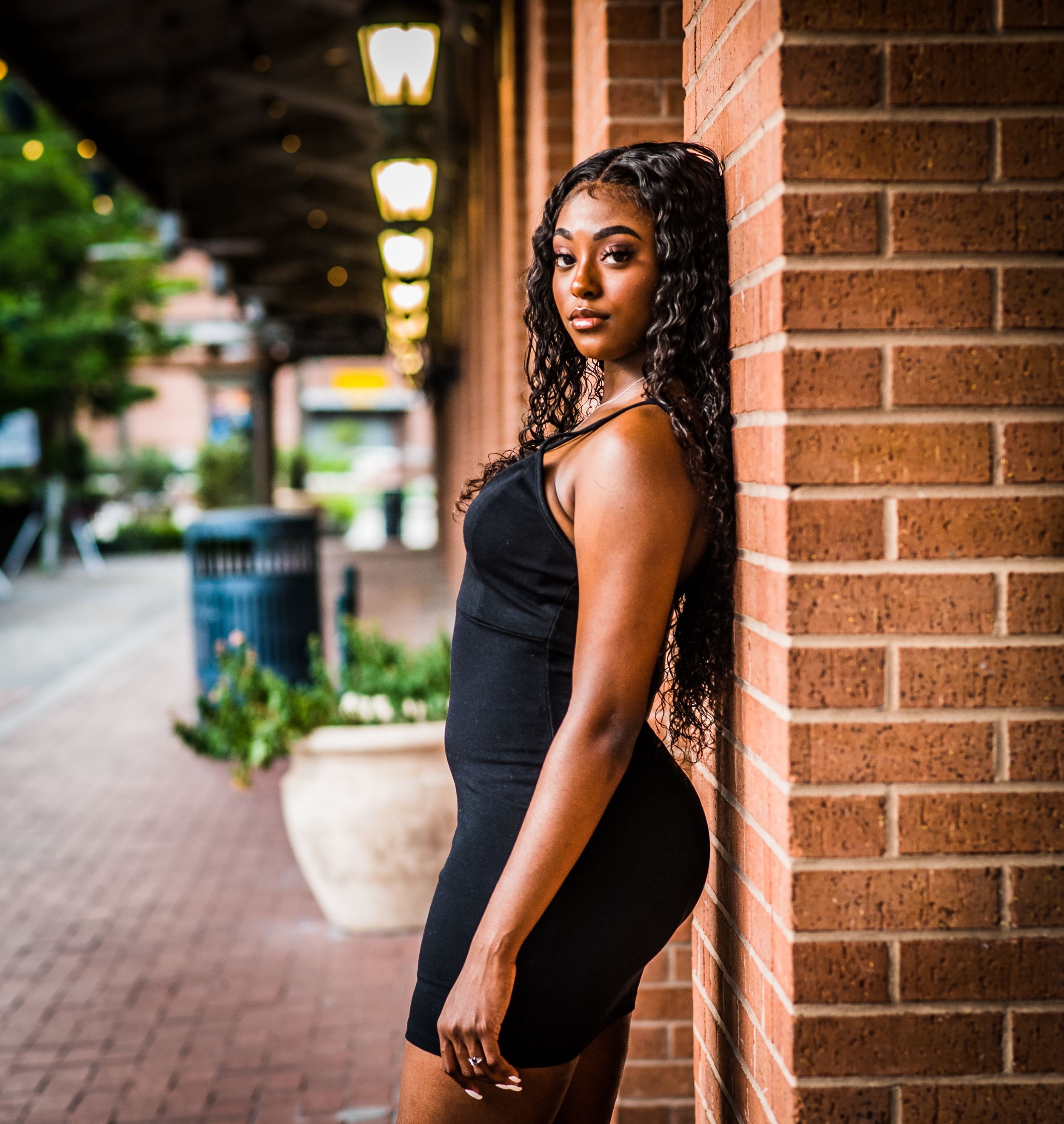 Elegant portrait of a model in black dress posing against brick wall with atmospheric evening lighting.