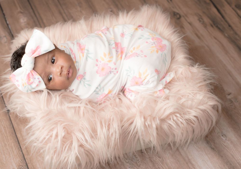 Newborn baby in floral swaddle and headband lying on pink furry backdrop.