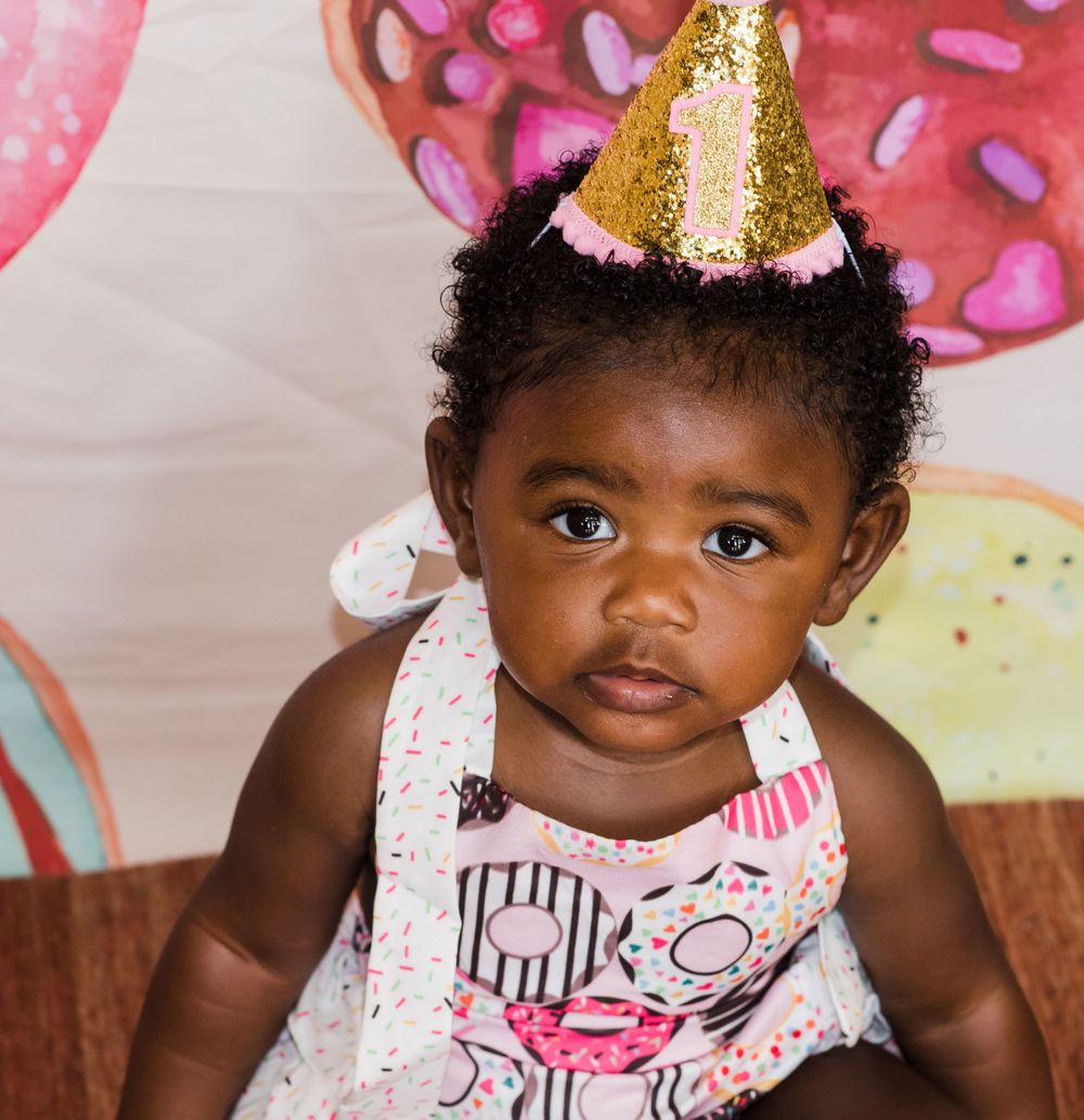 Baby in festive party hat and patterned dress celebrating during a donut-themed photoshoot.