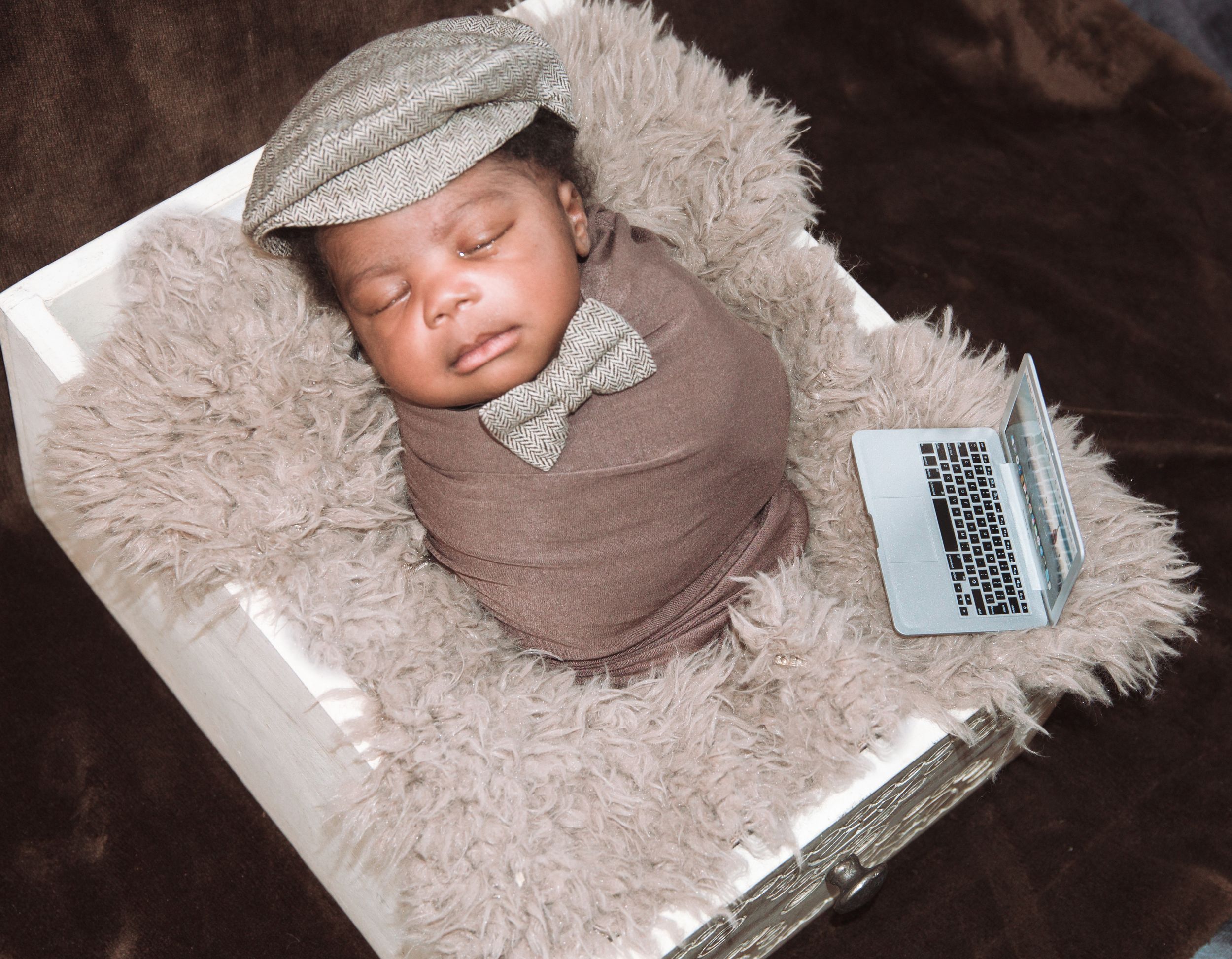 Newborn baby wrapped in brown swaddle sleeping on fluffy blanket next to laptop.