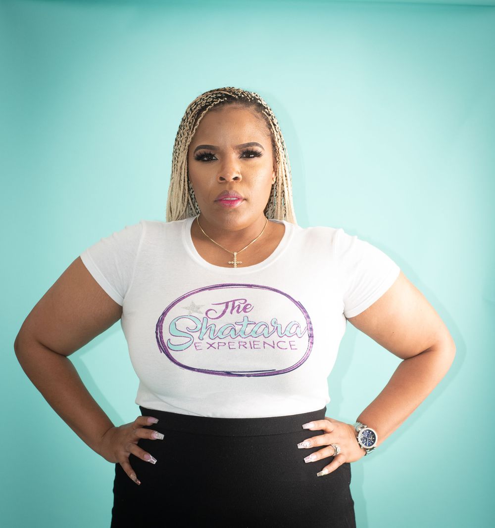 Casual brand photoshoot in white logo t-shirt and black pants against turquoise background.
