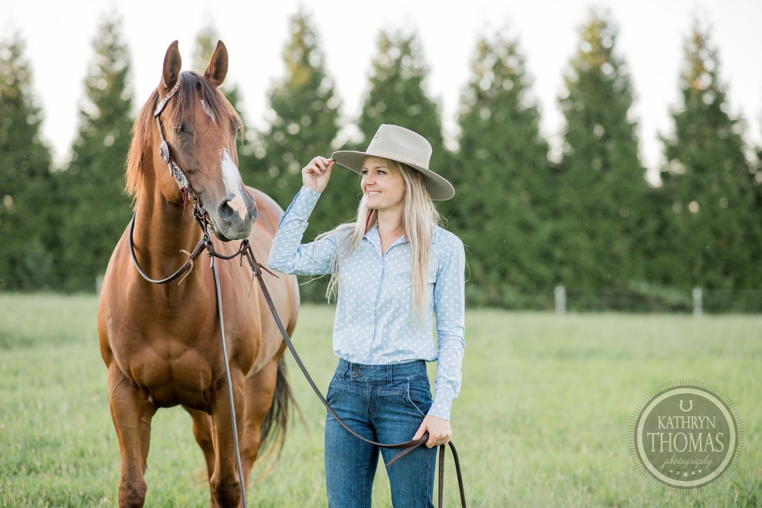 A chestnut horse with white markings stands beside someone in western wear near evergreen trees.