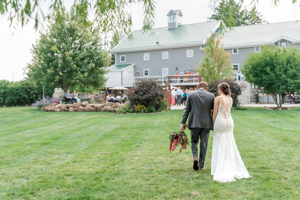 A newlywed couple walks with their dog across a rustic farm property with a barn in the background.