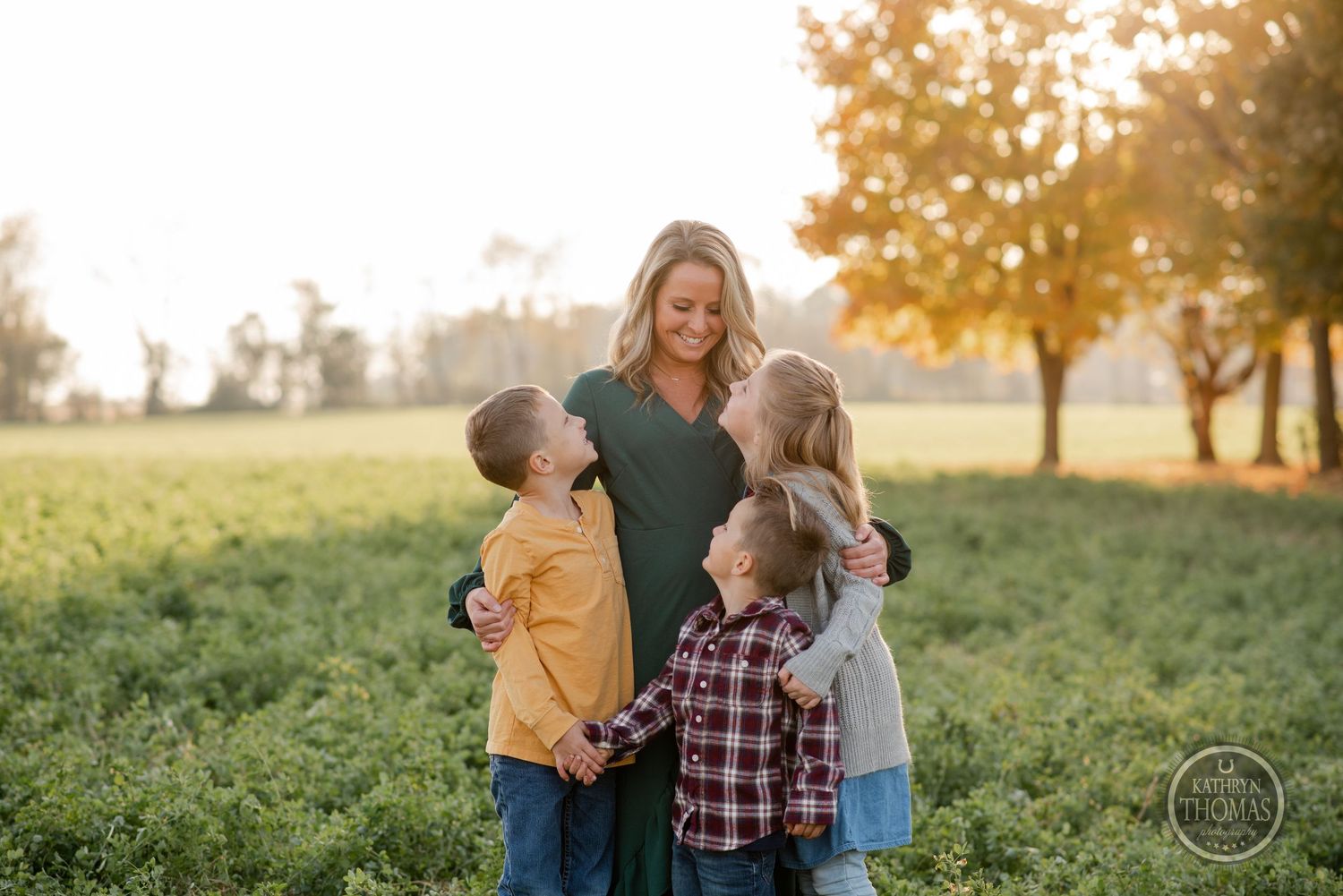 Family group huddled together in a green field during golden hour with autumn trees in background.