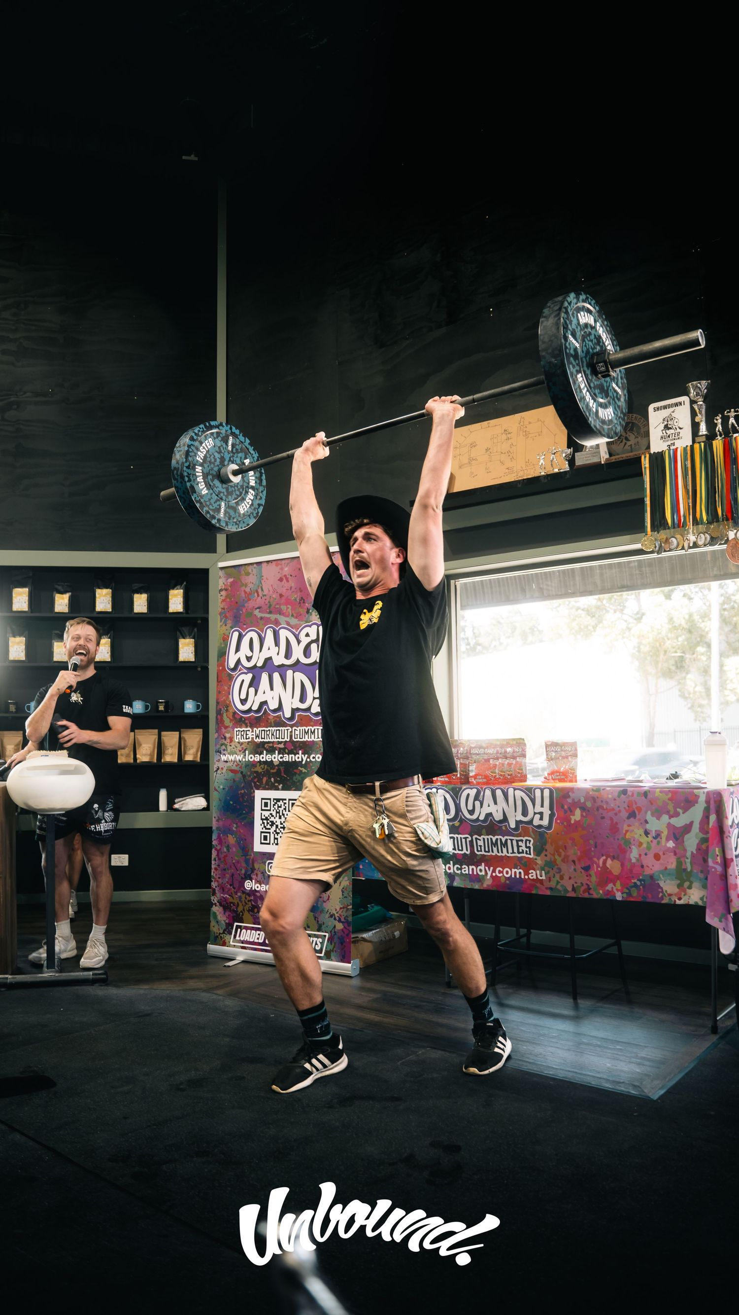 Athlete performs overhead press with barbell in dark gym setting with colorful backdrop.