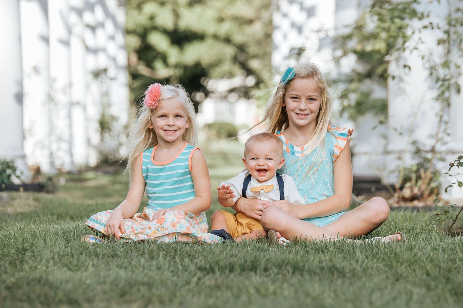 Three children in summer outfits sit together on grass on a sunny day.