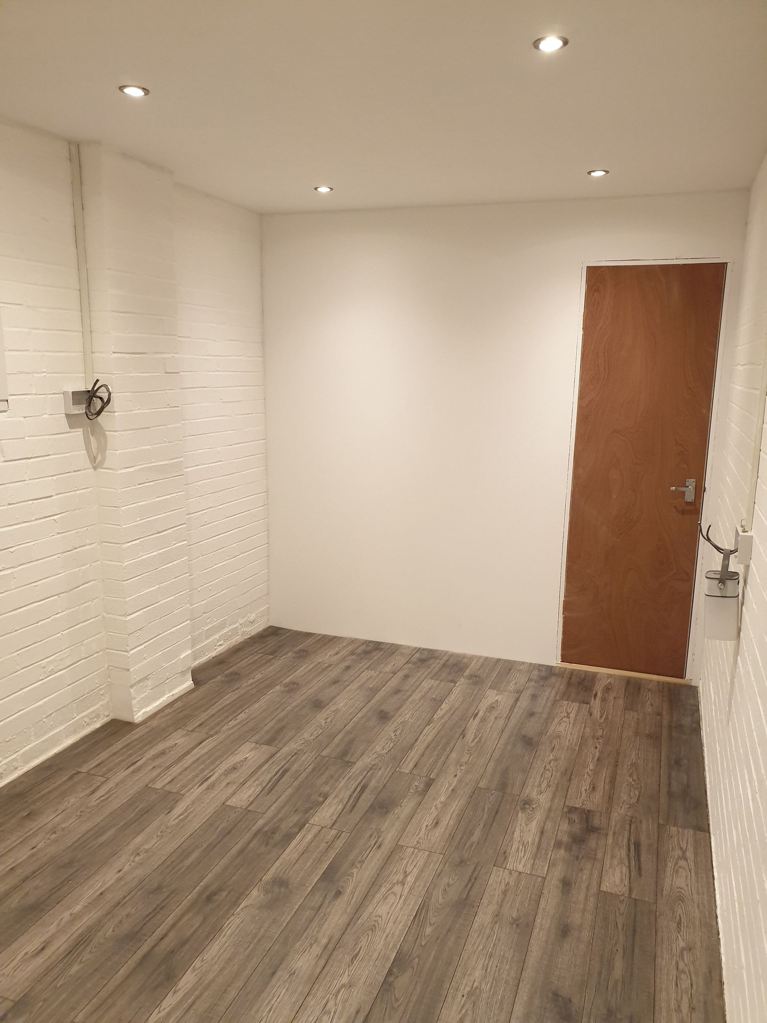 Newly renovated room with white painted brick walls, wooden floor and brown door.