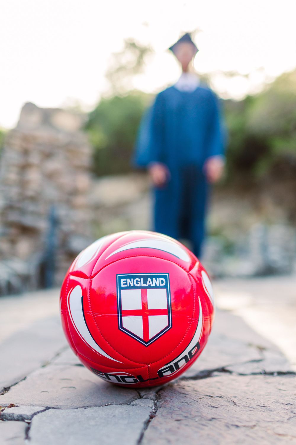 Red England soccer ball on stone pavement with graduate in blue gown in background.