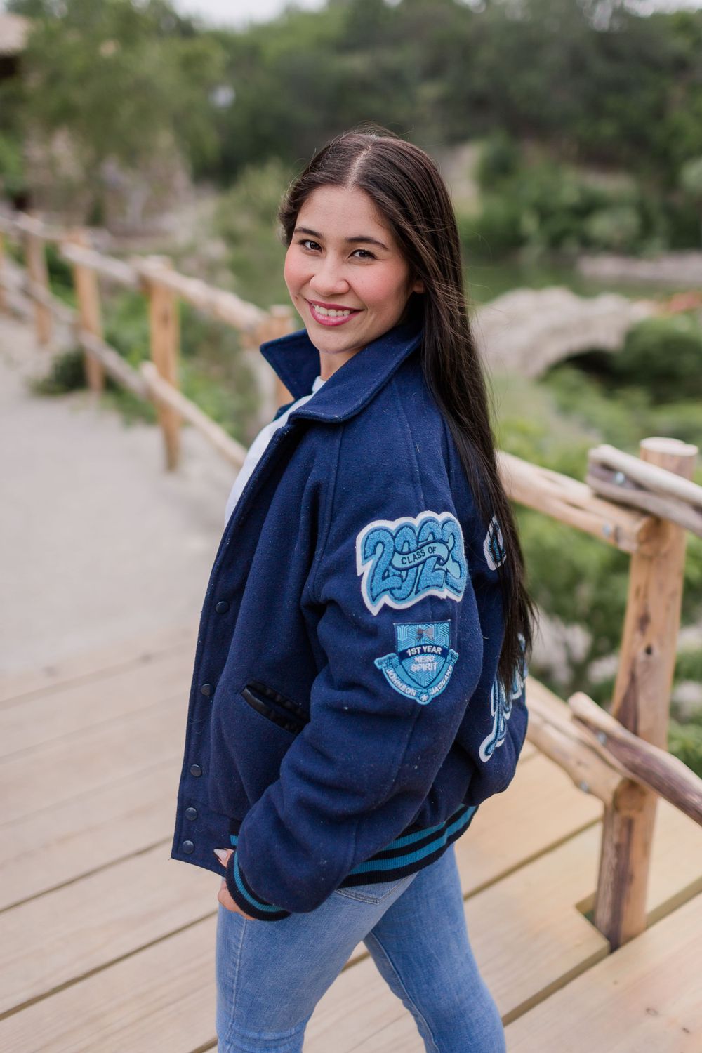 A person in a varsity jacket poses on a wooden bridge path in a natural setting.