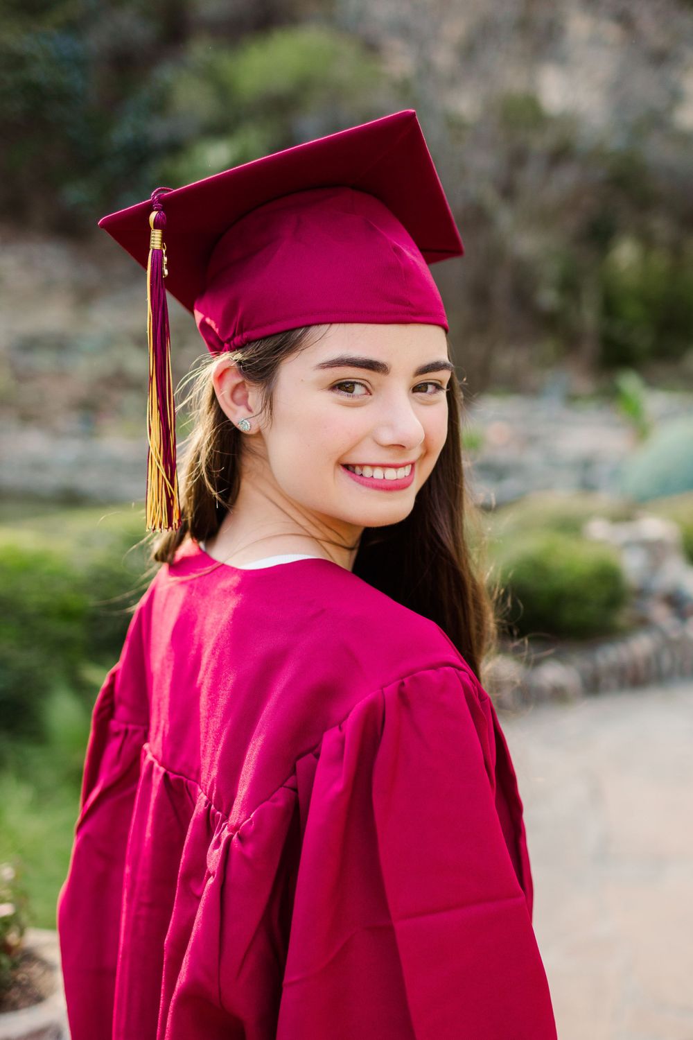 Graduate in red cap and gown poses against stone backdrop.