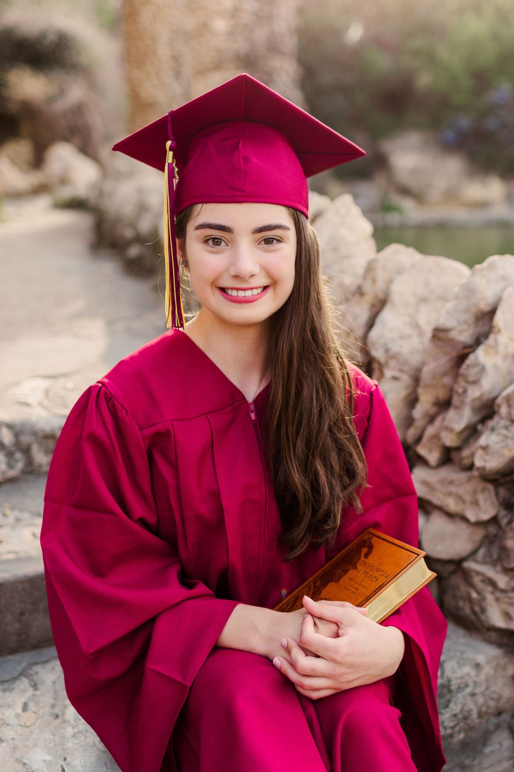 Graduate wearing crimson cap and gown holding book against stone wall.