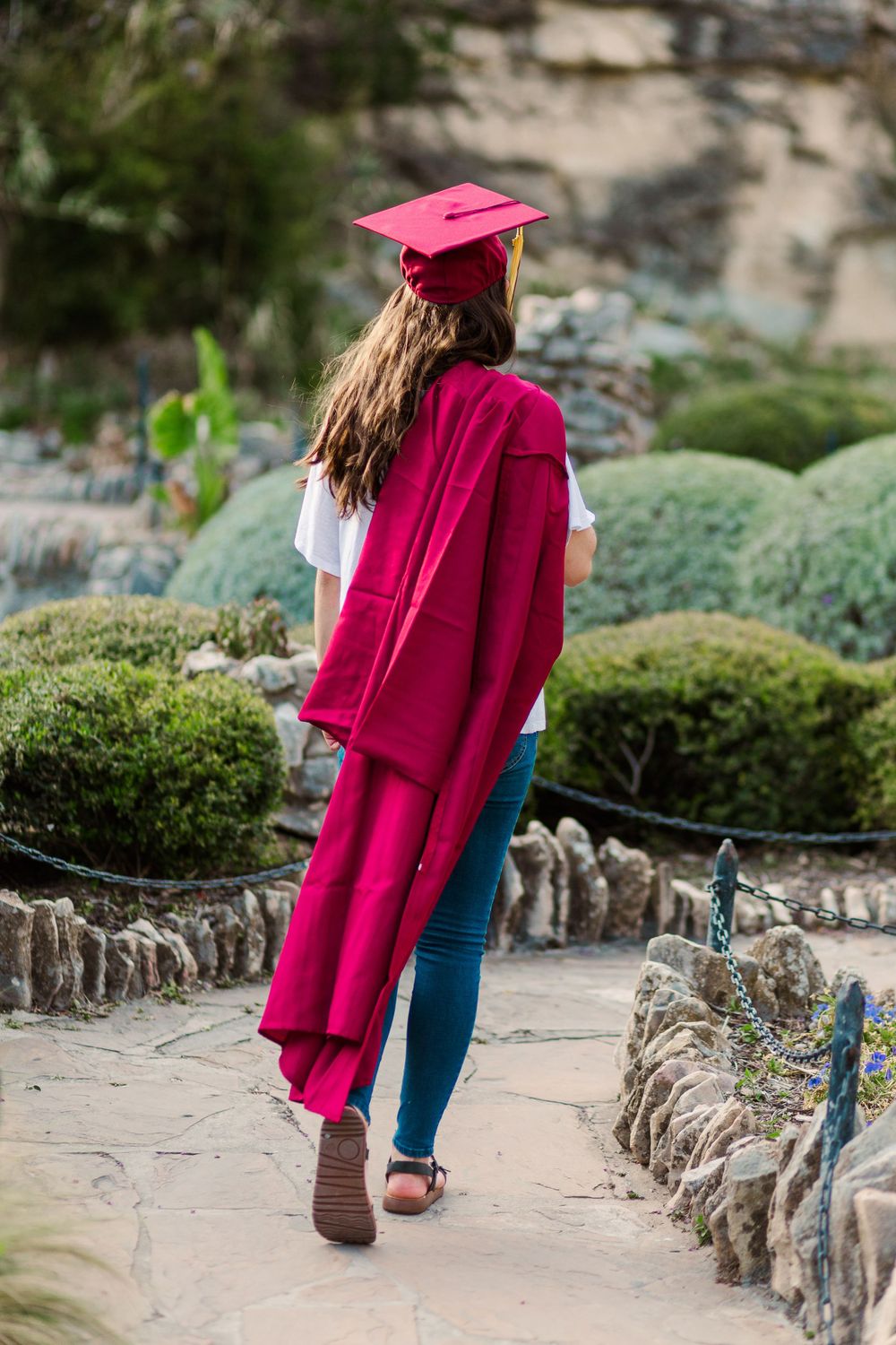 Graduate in burgundy gown walking along garden path with stone wall.