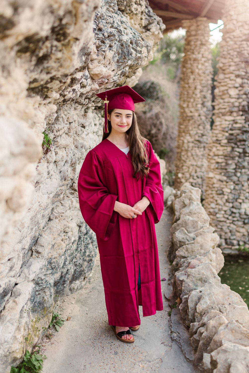 Graduate in crimson regalia standing on stone path near rock wall.