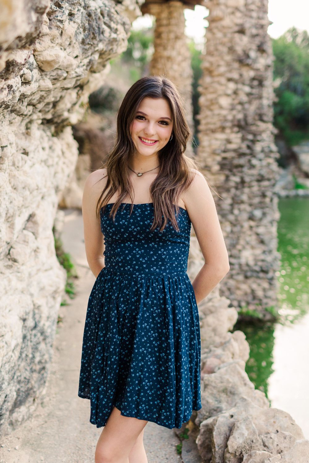 Young woman in a navy blue polka dot dress poses against stone columns in an outdoor setting.