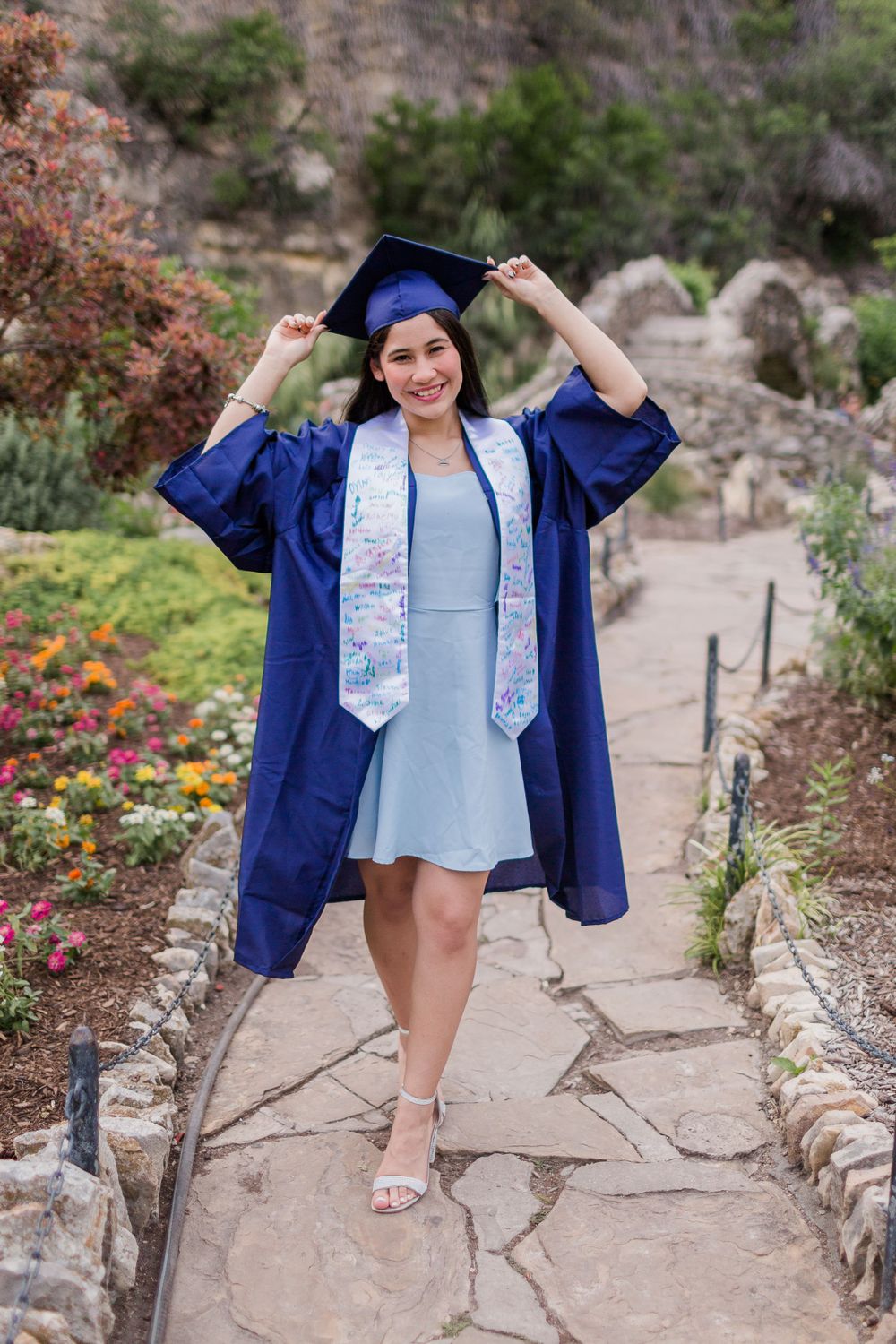 A graduate in blue cap and gown poses on a stone path surrounded by flowers.