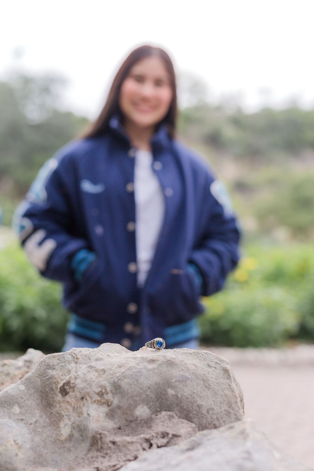 A class ring sits on a rock with a blurred figure wearing a letterman jacket in the background.