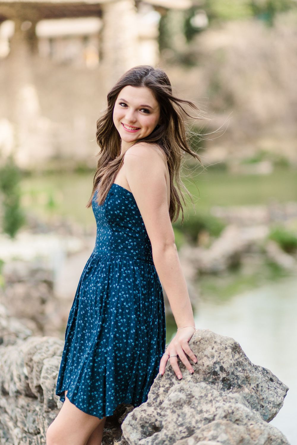 Woman in blue patterned dress poses on rocky outcrop near water.