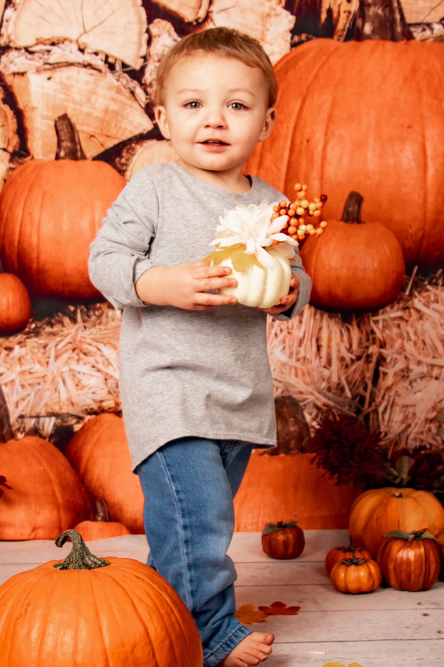 A child in grey holds a small gift while standing among orange pumpkins and autumn decorations.