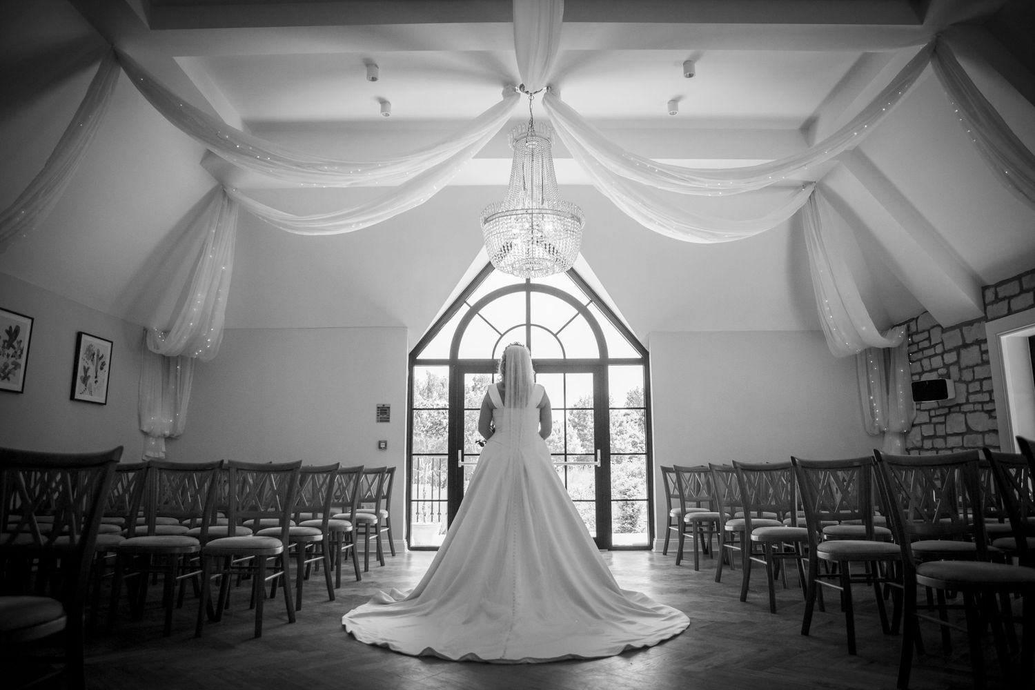 Elegant bride in flowing white gown stands alone in beautifully draped wedding ceremony hall, black and white.