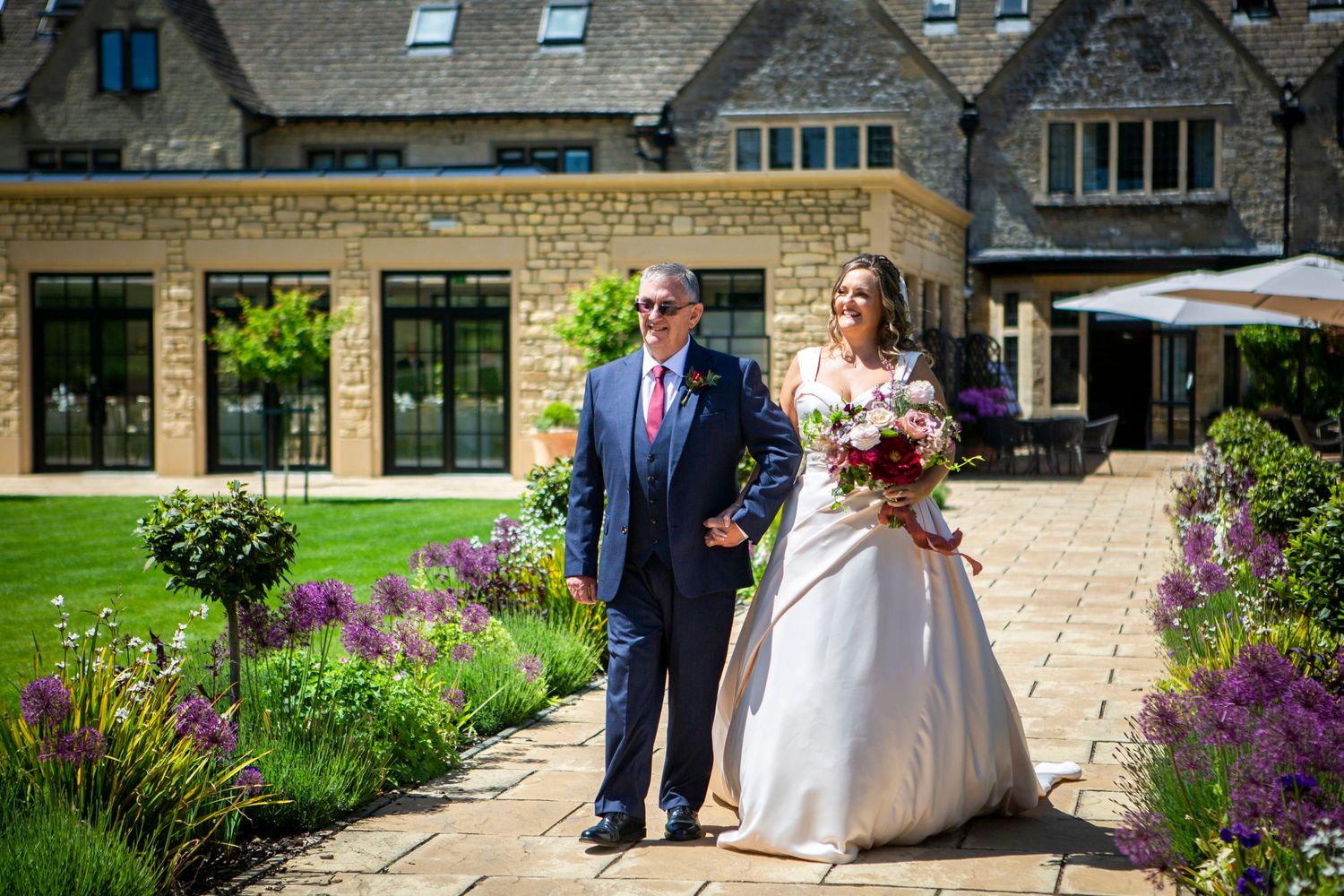 Bride holding colorful bouquet walks with suited man through stunning purple flower garden at venue.