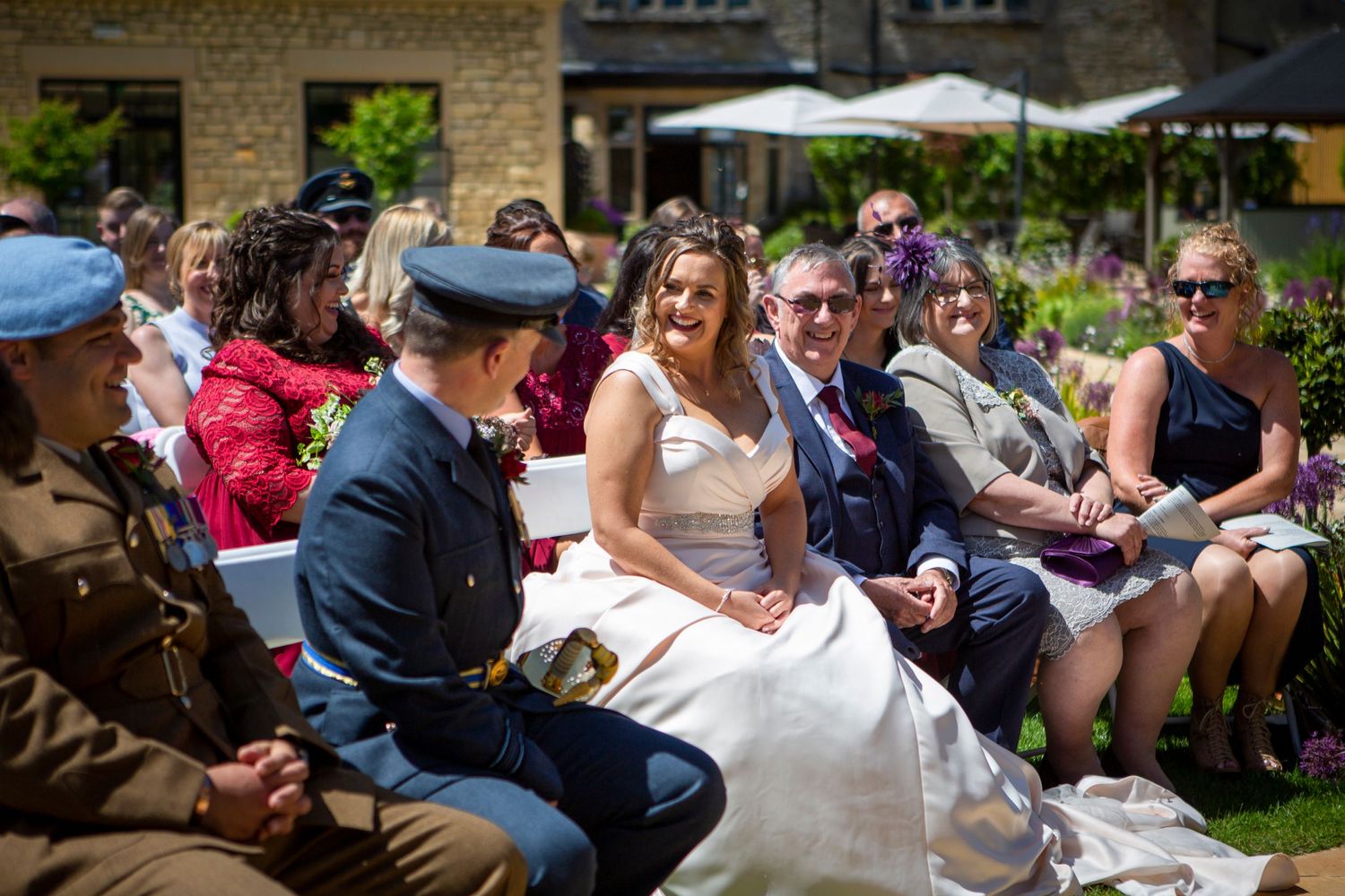 Bride smiling at groom in military uniform during outdoor wedding ceremony with seated guests behind.
