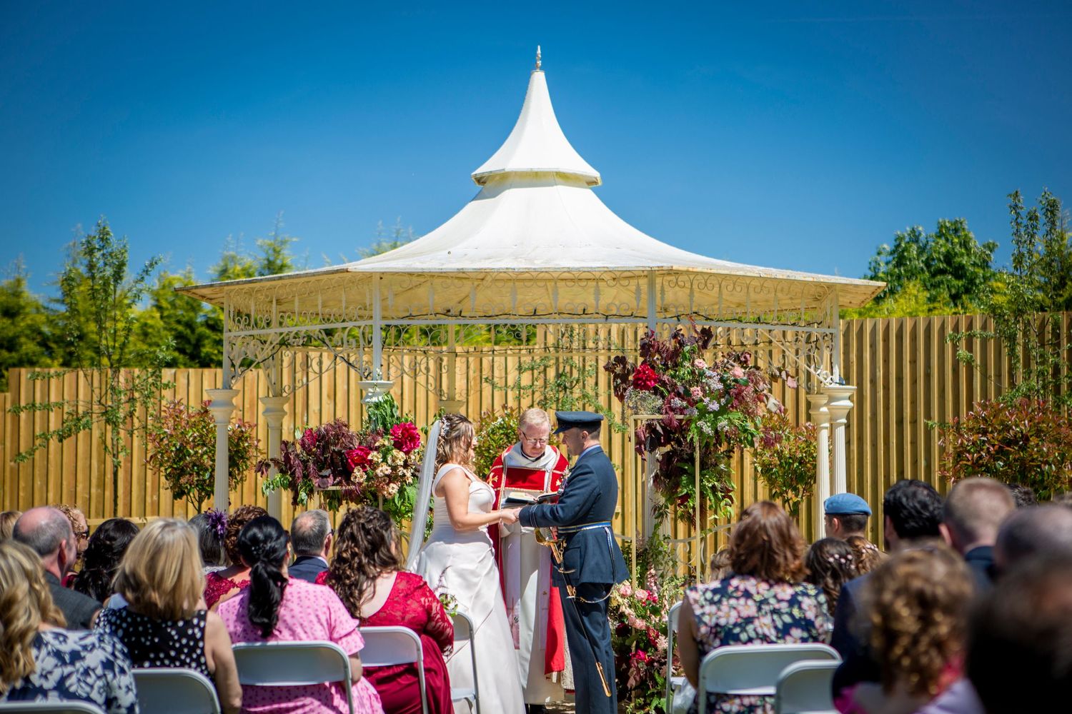 Outdoor wedding ceremony with couple at altar beneath white pavilion on a sunny blue sky day.