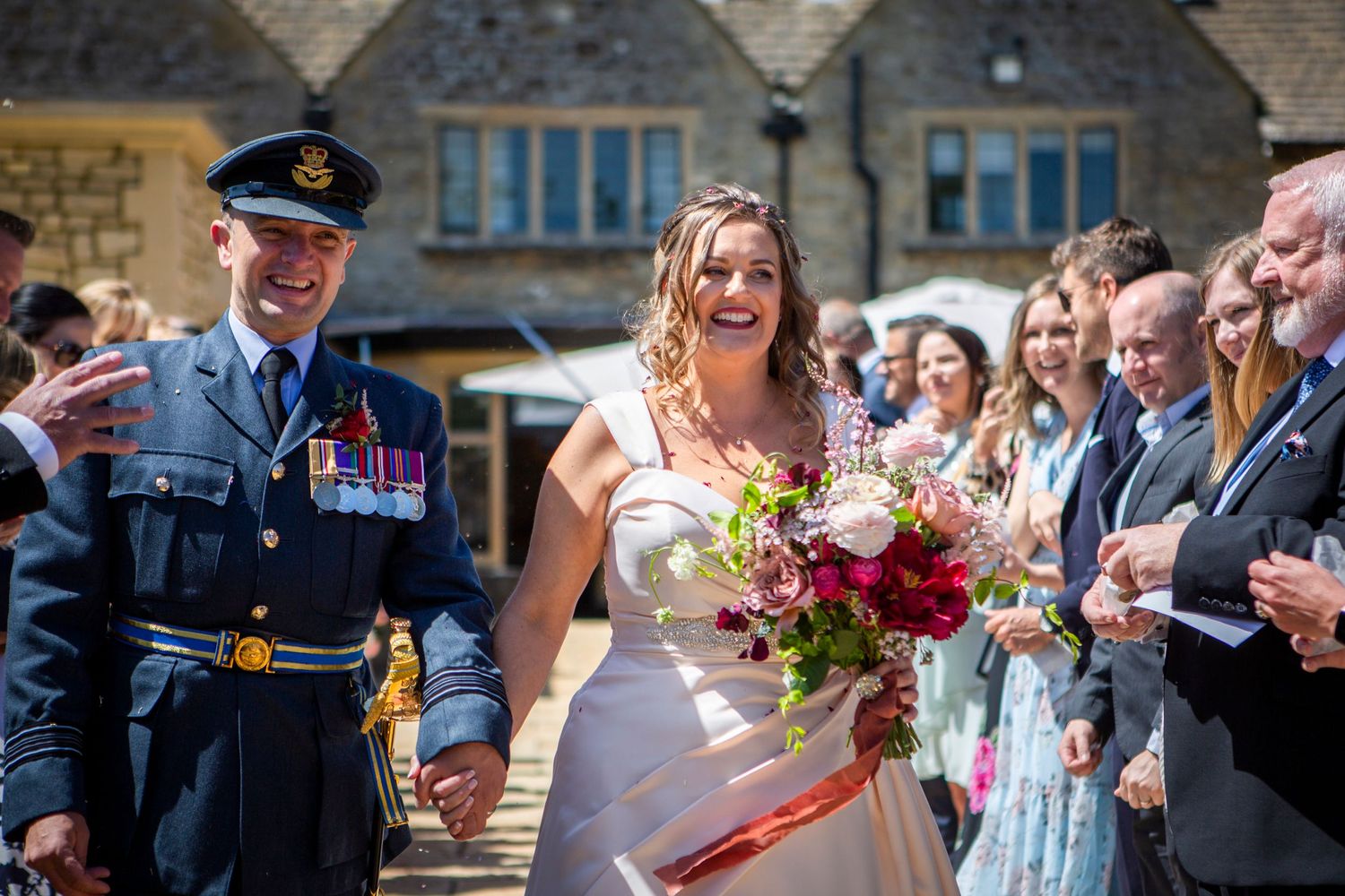 Bride laughing with groom in RAF uniform walking hand in hand through confetti-throwing wedding guests.