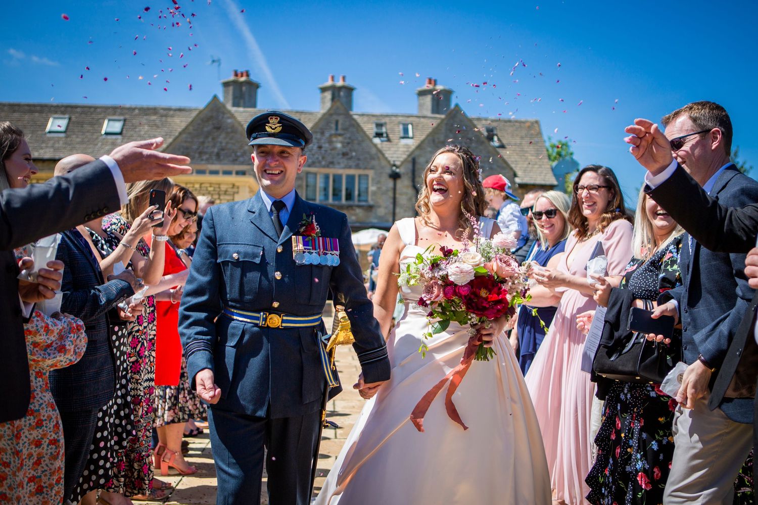 Smiling bride and groom in military uniform walk through joyful confetti shower after wedding ceremony.