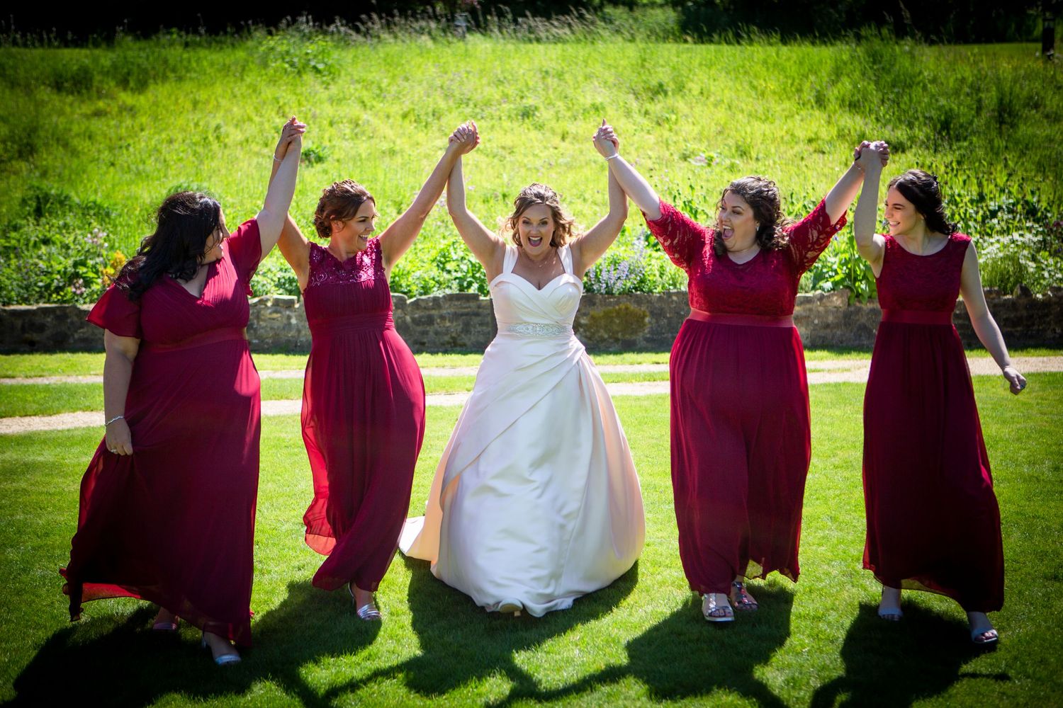 Bride in white gown celebrates with four bridesmaids in red dresses raising hands on green lawn.