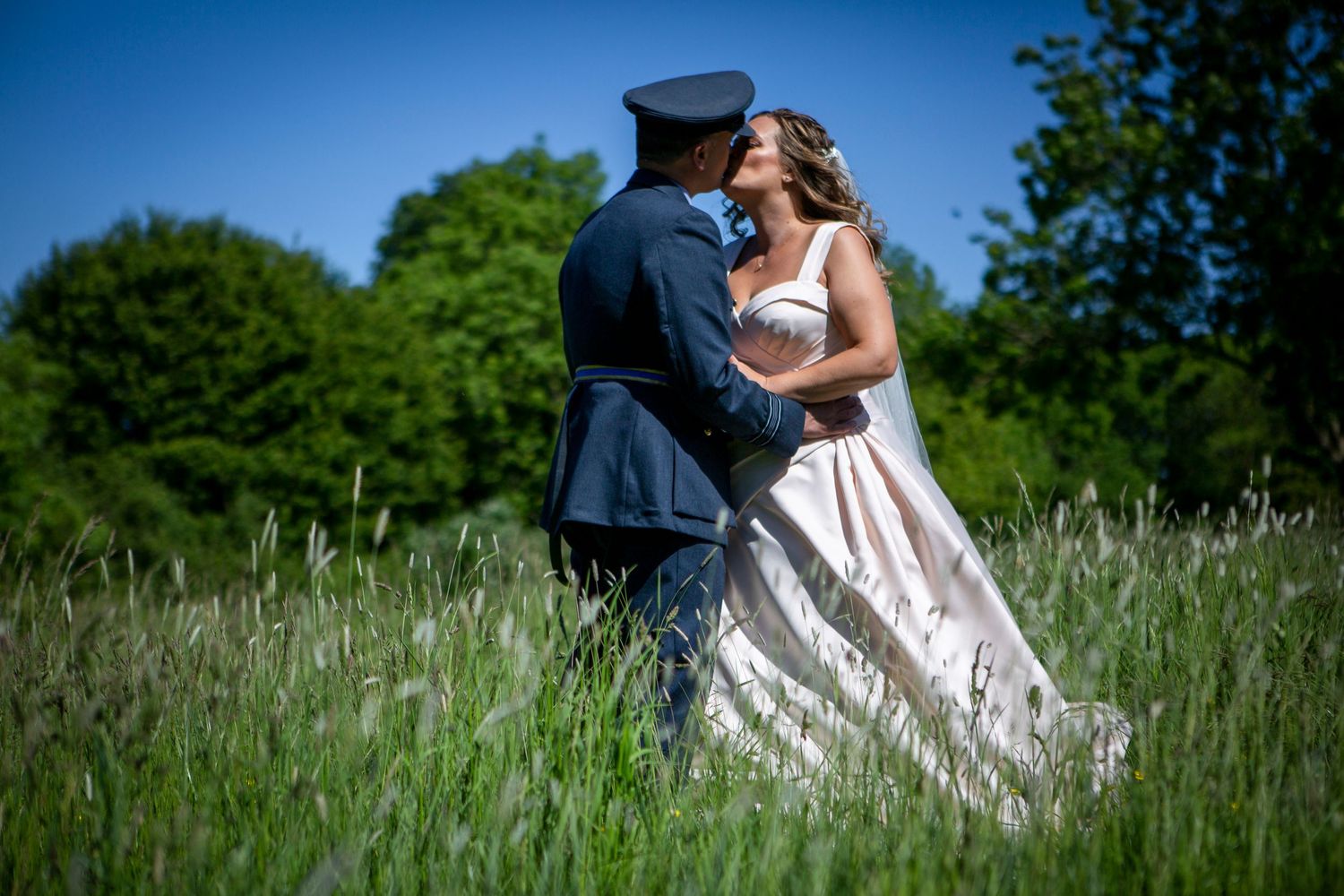 Bride and groom in military uniform share romantic kiss embracing in tall green meadow grass.