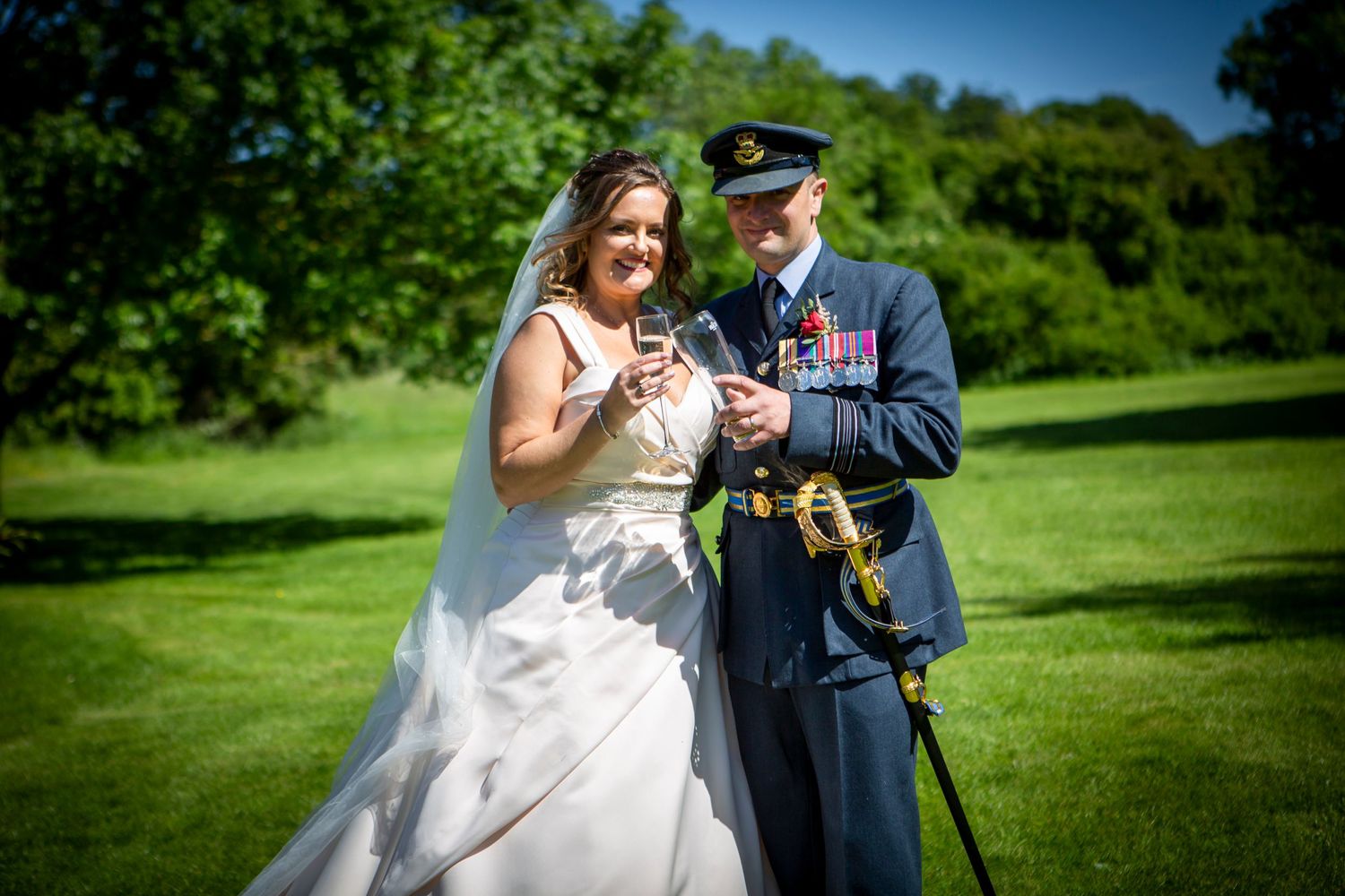 Bride and groom in RAF dress uniform clink champagne glasses while smiling on lush green lawn.