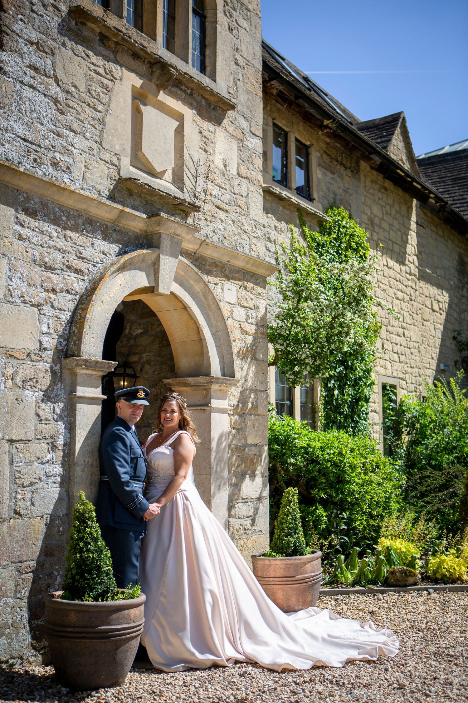 Bride and groom in military uniform pose together at arched doorway of historic stone manor house.