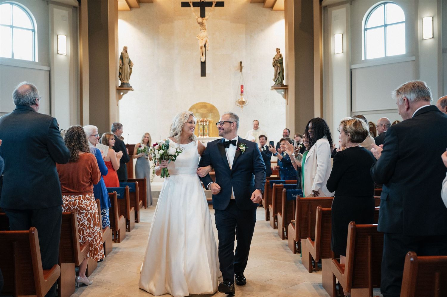 A couple walks down the church aisle while guests stand and celebrate.