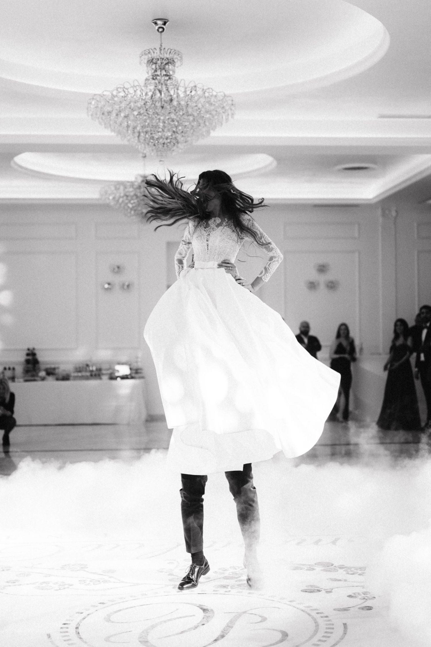 A black and white photo of a spinning bride in a flowing white dress under an elegant crystal chandelier.