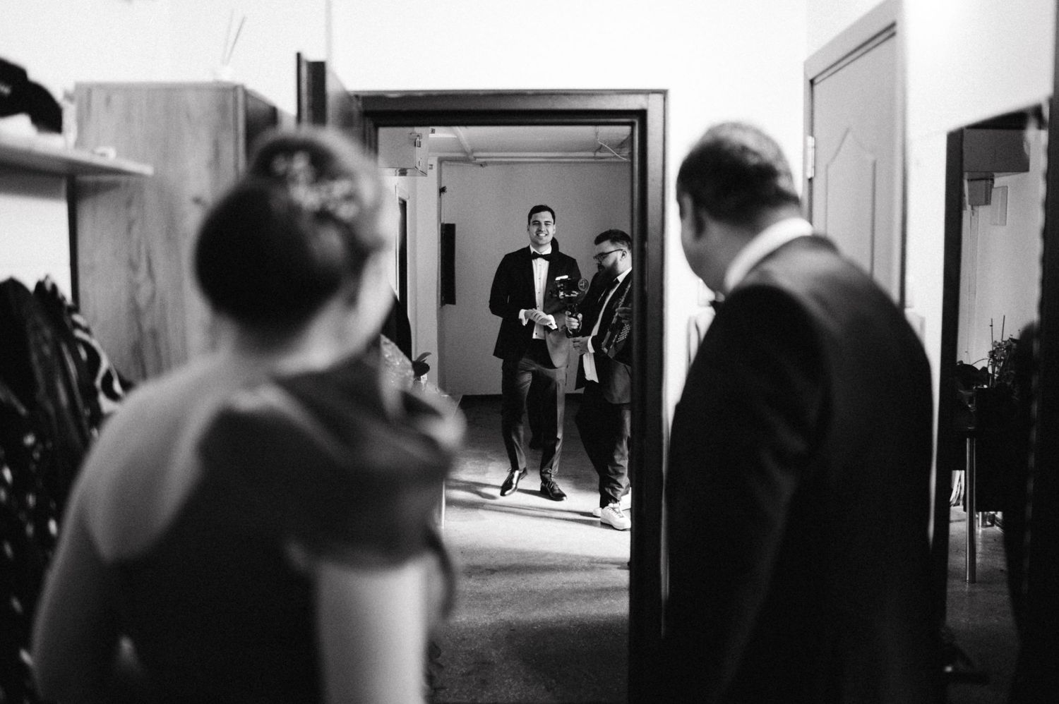 Black and white candid shot of wedding guests in formal attire gathered in a hallway.